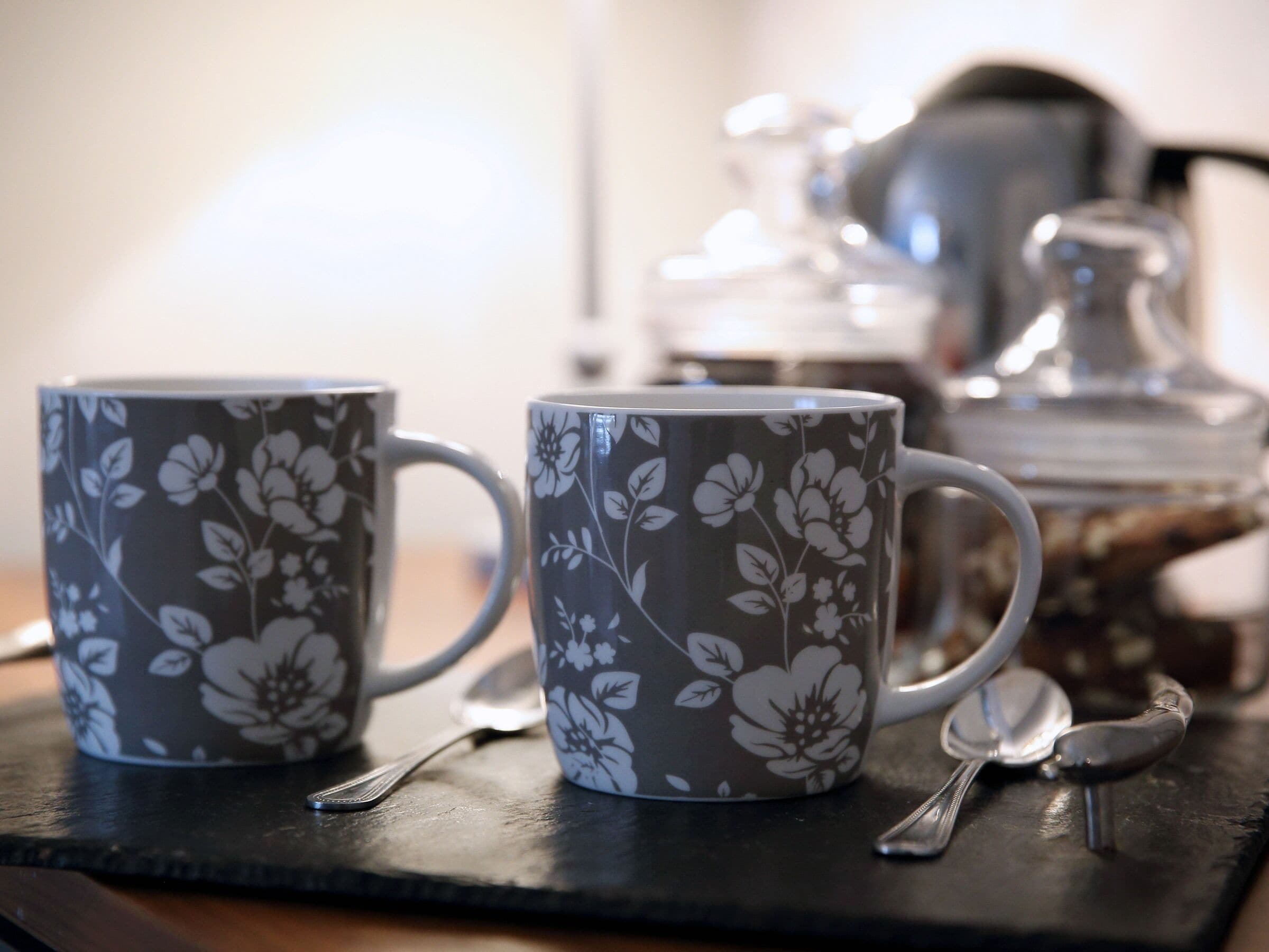 Dos tazas con estampado floral sobre una bandeja de pizarra oscura, con una cuchara y un hervidor de agua al fondo.