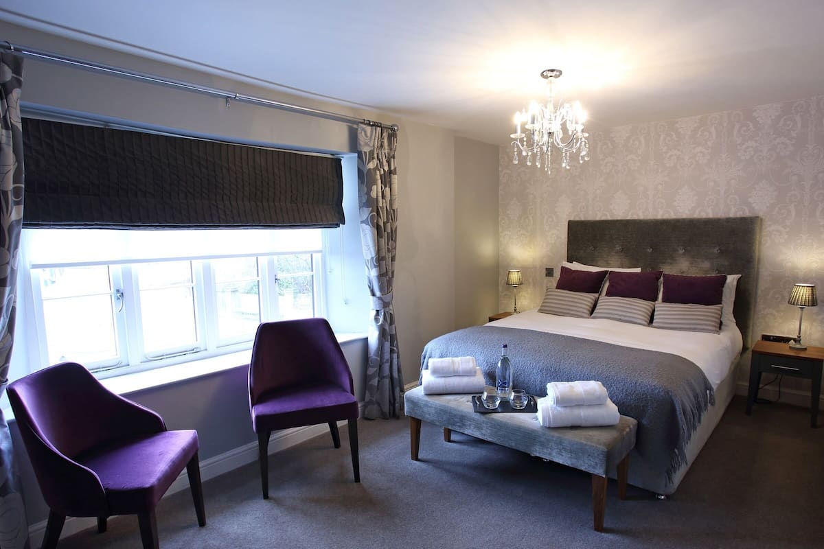 A hotel room with a large bed, two chairs, a bedside table with a lamp, and a window with curtains.