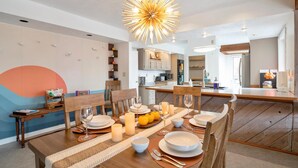 Un coin repas avec une table en bois dressée pour le repas, un lustre moderne et une cuisine en arrière-plan.