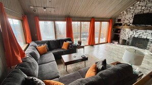 Un salon chaleureux avec un canapé d’angle, une cheminée et de grandes fenêtres ornées de rideaux orange.