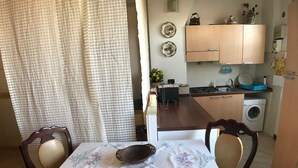 A dining area with a table set for two, a kitchen in the background with wooden cabinets and a washing machine, and a curtain partially covering a window. - Porta Camollia Apartment (Siena)