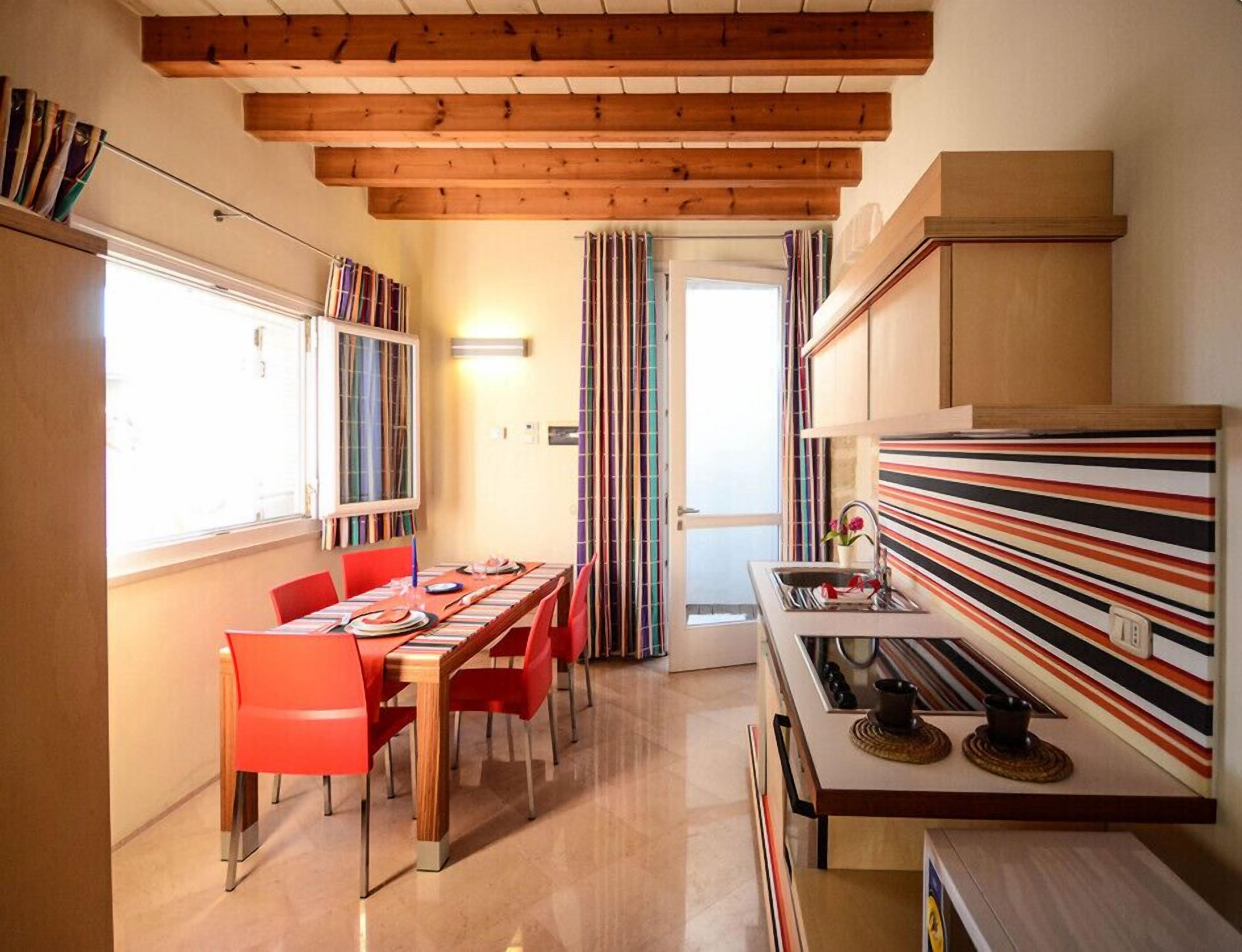 A compact kitchen with a striped backsplash, a dining area with red chairs, and a wooden ceiling.