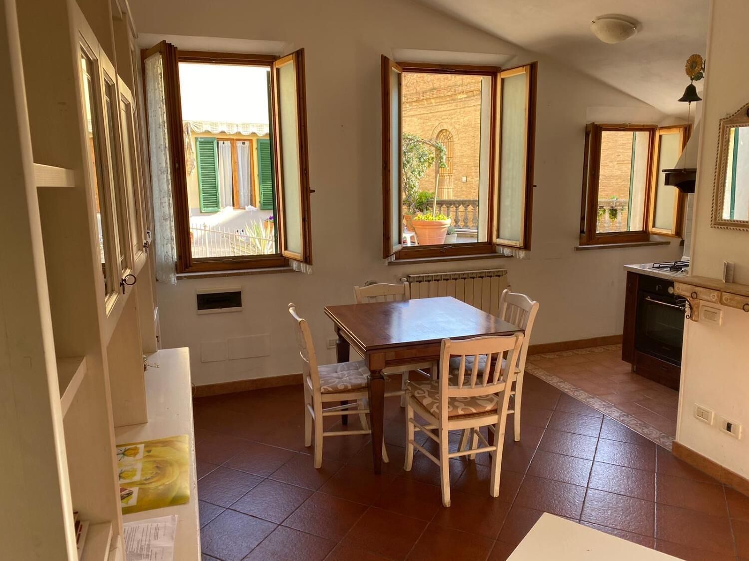 A dining area with a wooden table and chairs, a window with a view of a building and greenery, and a kitchen area with a stove and cabinets.