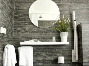 A bathroom with a dark brick wall, a round mirror, a white shelf with toiletries, a potted plant, a towel rack, and a toilet paper holder. - Akkeri Guesthouse (Stykkisholmur)