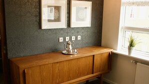 A wooden console table with a silver pitcher, framed pictures on the wall, and a window with a plant. - Akkeri Guesthouse (Stykkisholmur)