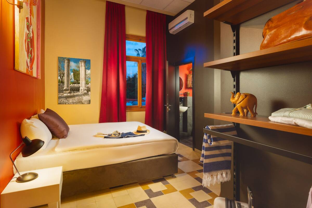 A hotel room with a bed, a desk, a shelf with a statue, and a window with red curtains.