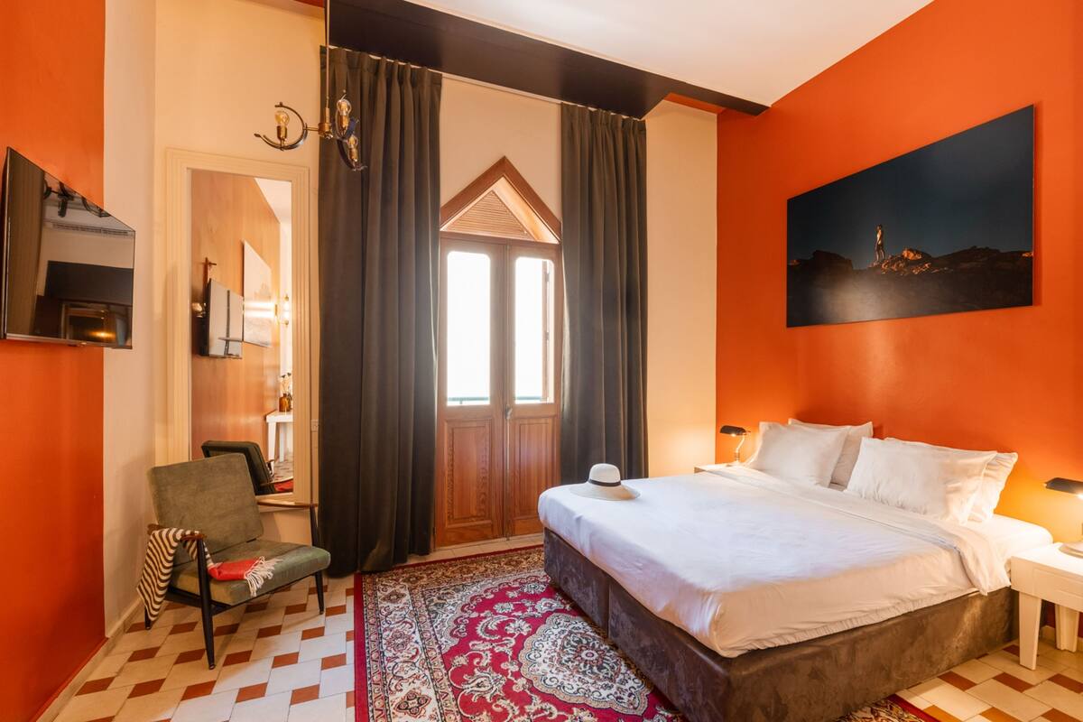 A hotel room with a bed, a chair, a desk, a mirror, a painting, and a window with curtains.