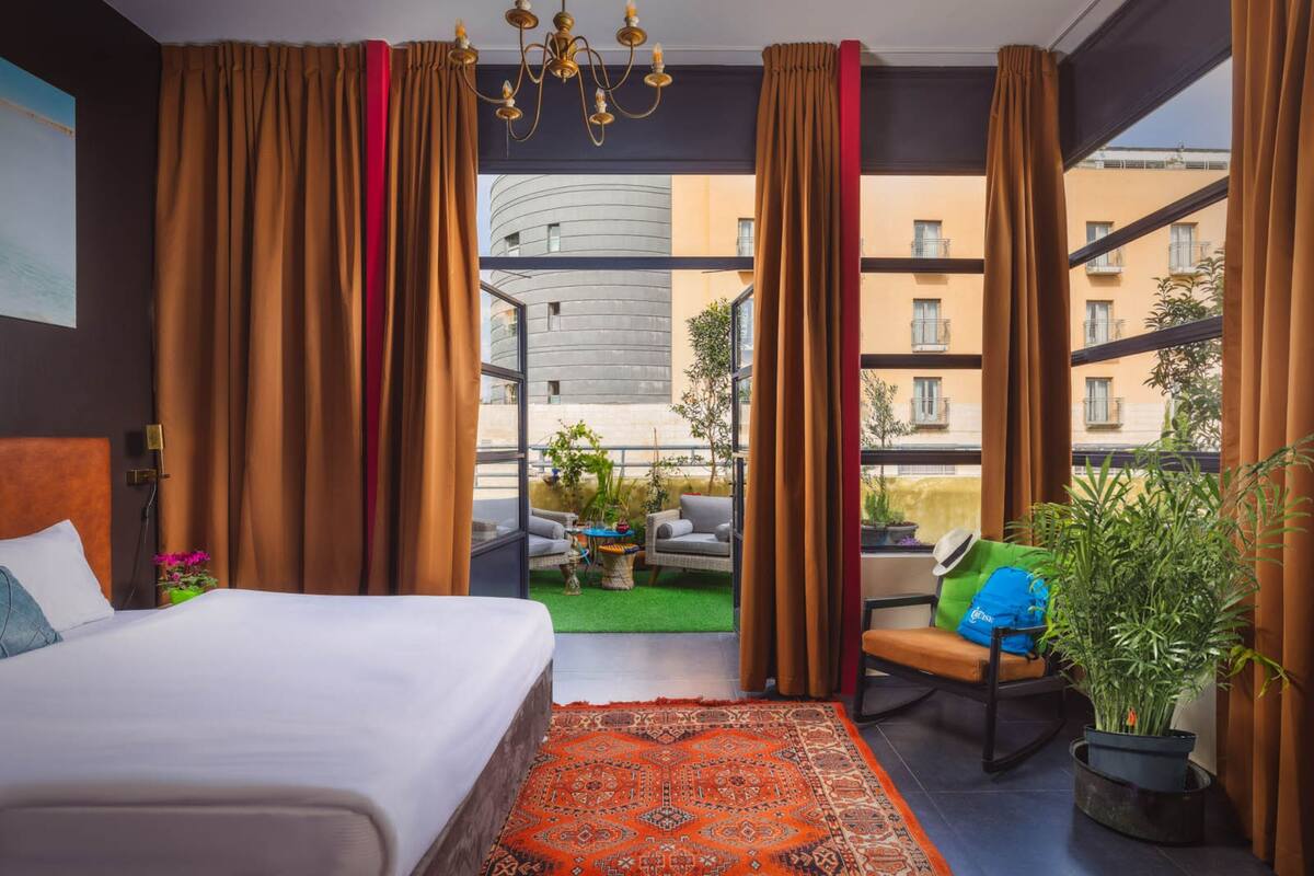A modern hotel room with a large bed, a balcony with outdoor seating, and a view of the cityscape.