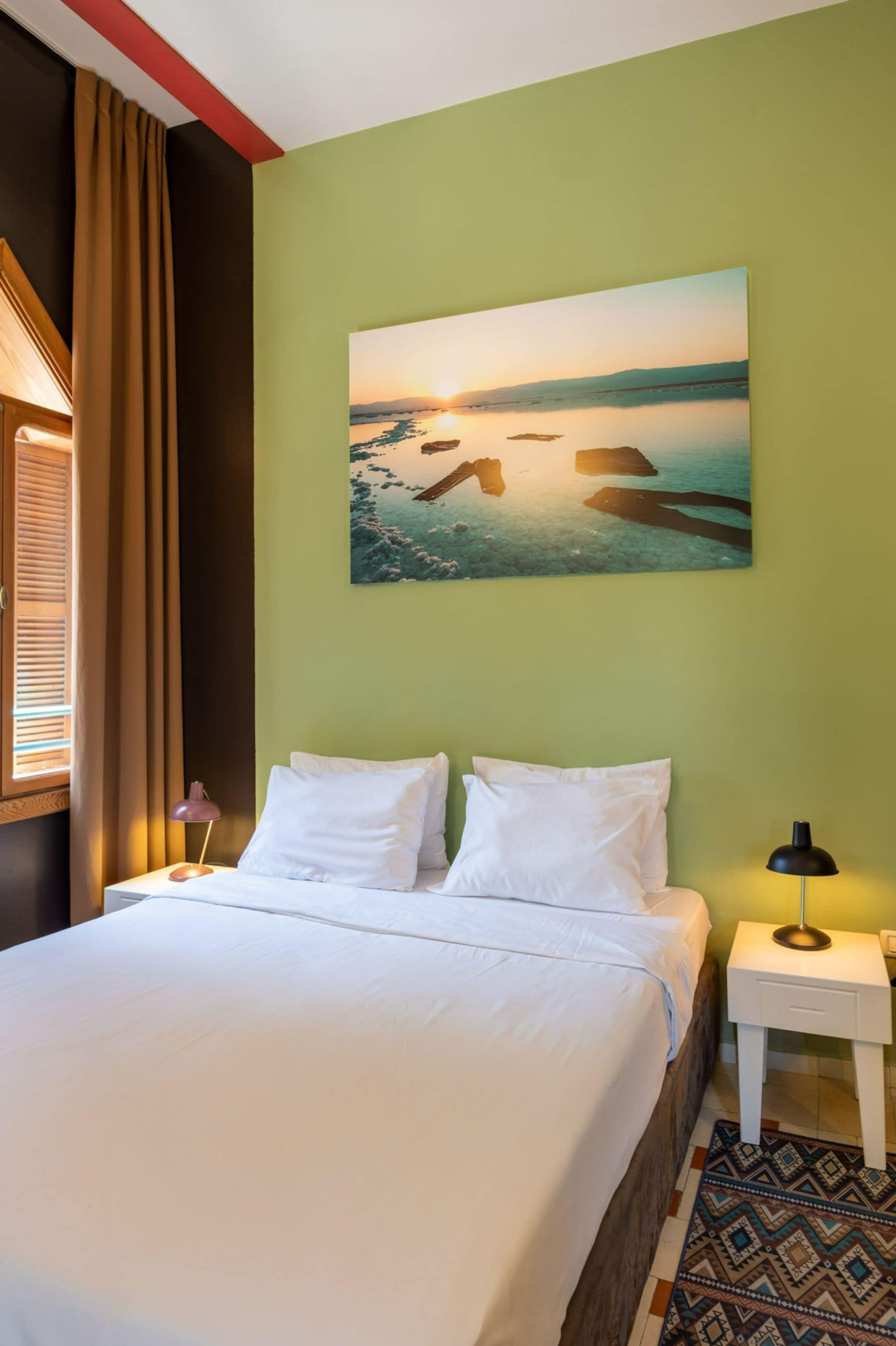 A neatly made bed with white linens, a painting of a sunset over water on the wall, and a patterned rug on the floor.
