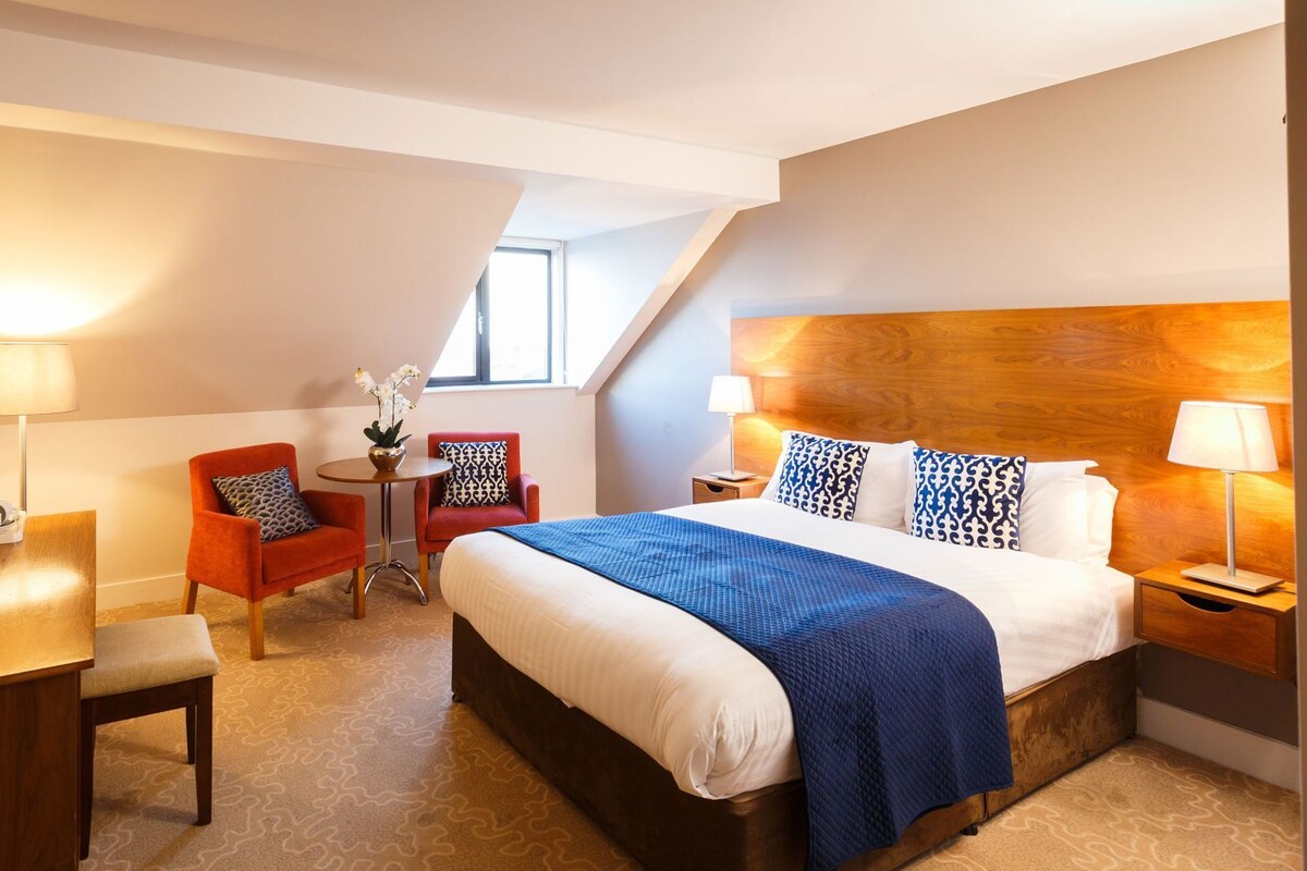 A hotel room with a bed, bedside tables, a desk with a telephone, a chair, a small table with a plant, and a window.