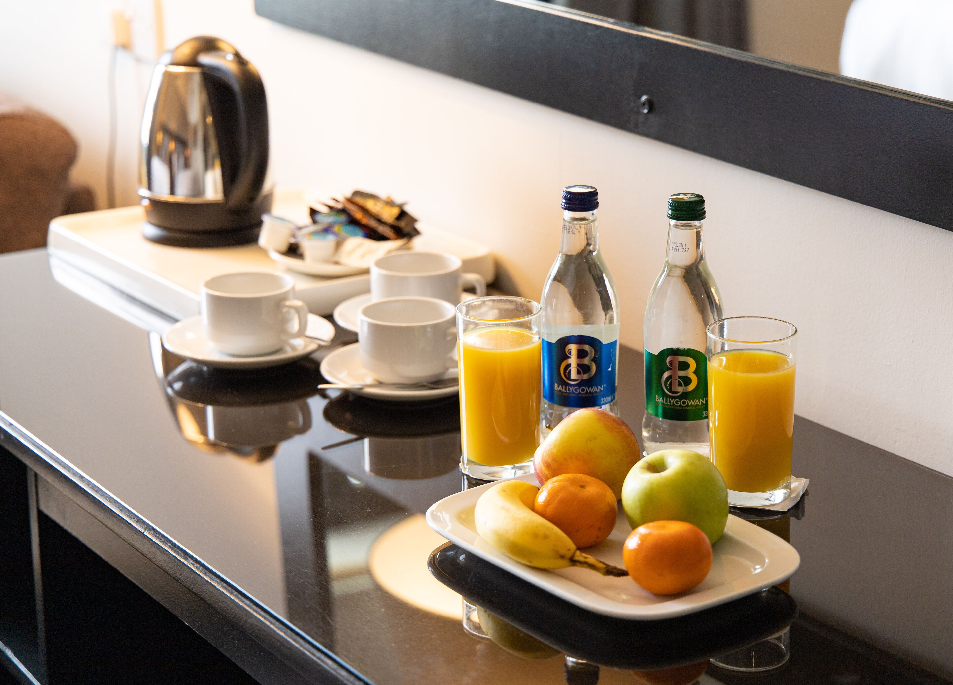 Un desayuno con un plato de frutas, dos botellas de jugo y varias tazas y platitos sobre una superficie reflectante.