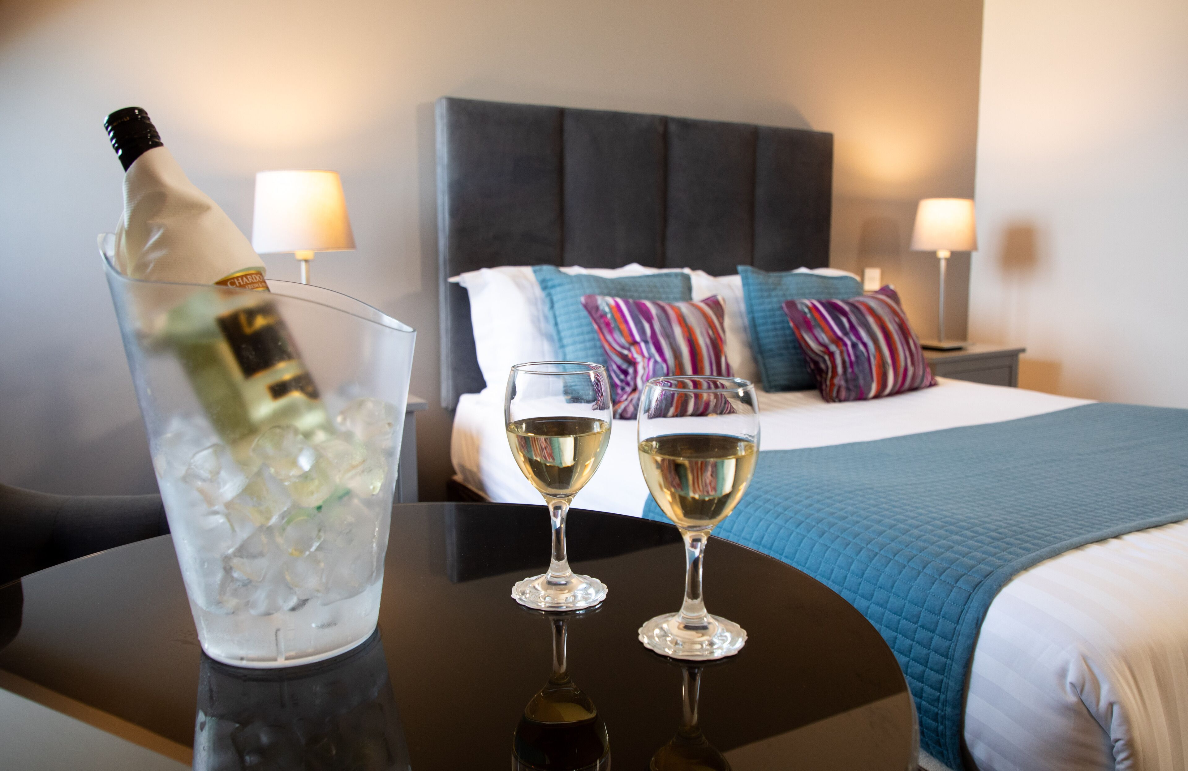 A hotel room with a bed, two bedside lamps, a bedside table with a bottle of champagne and two glasses of wine.