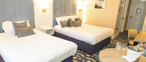 A hotel room with two beds, a small table with glasses and a bottle, and a door in the background.