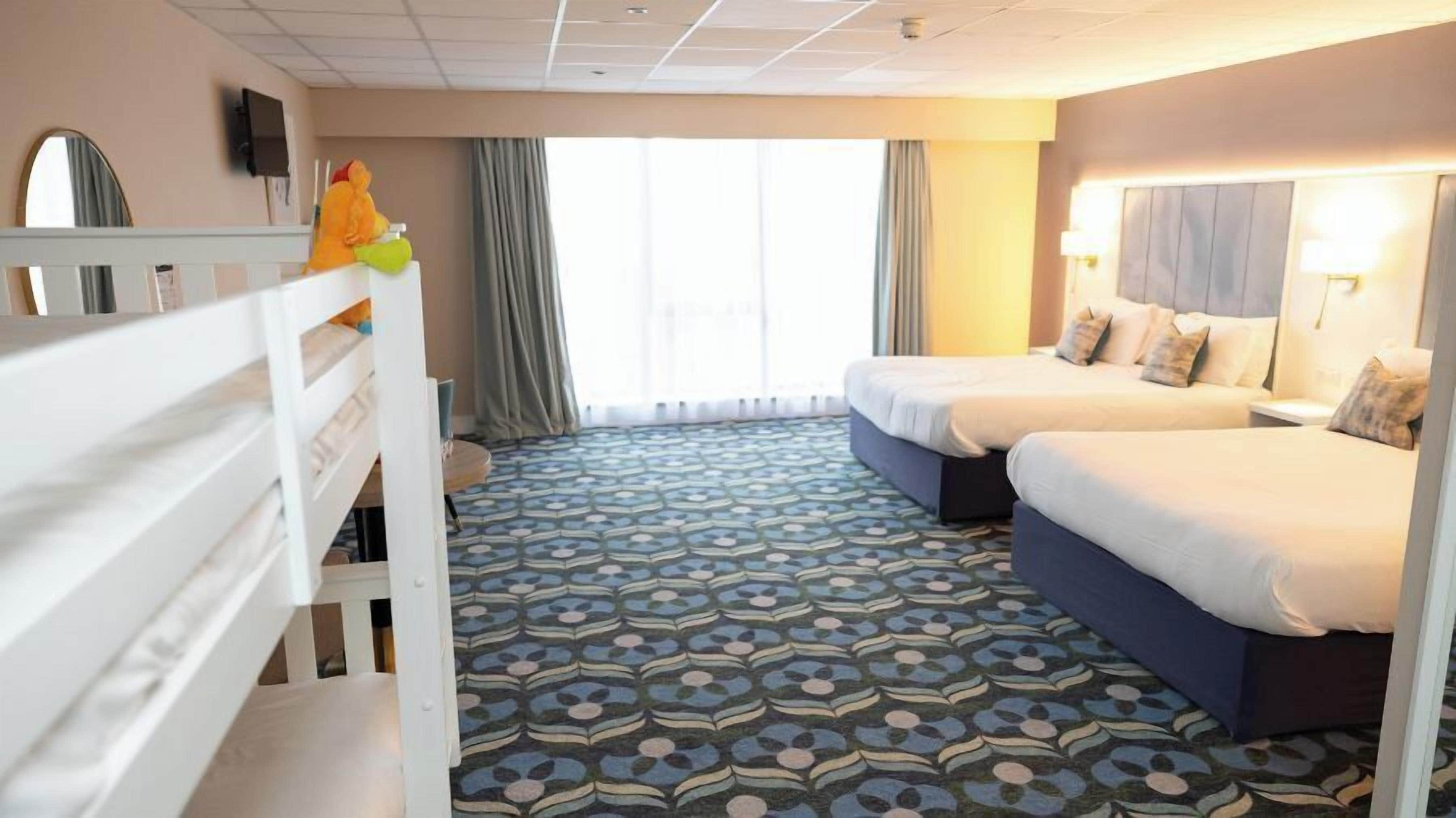 A hotel room with a patterned carpet, two beds, a small table, a mirror, and a window with curtains.