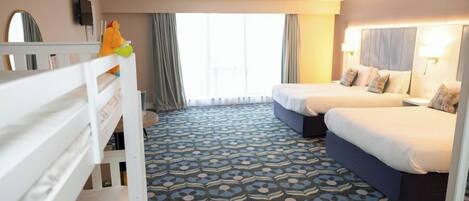 A hotel room with a patterned carpet, two beds, a small table, a mirror, and a window with curtains.