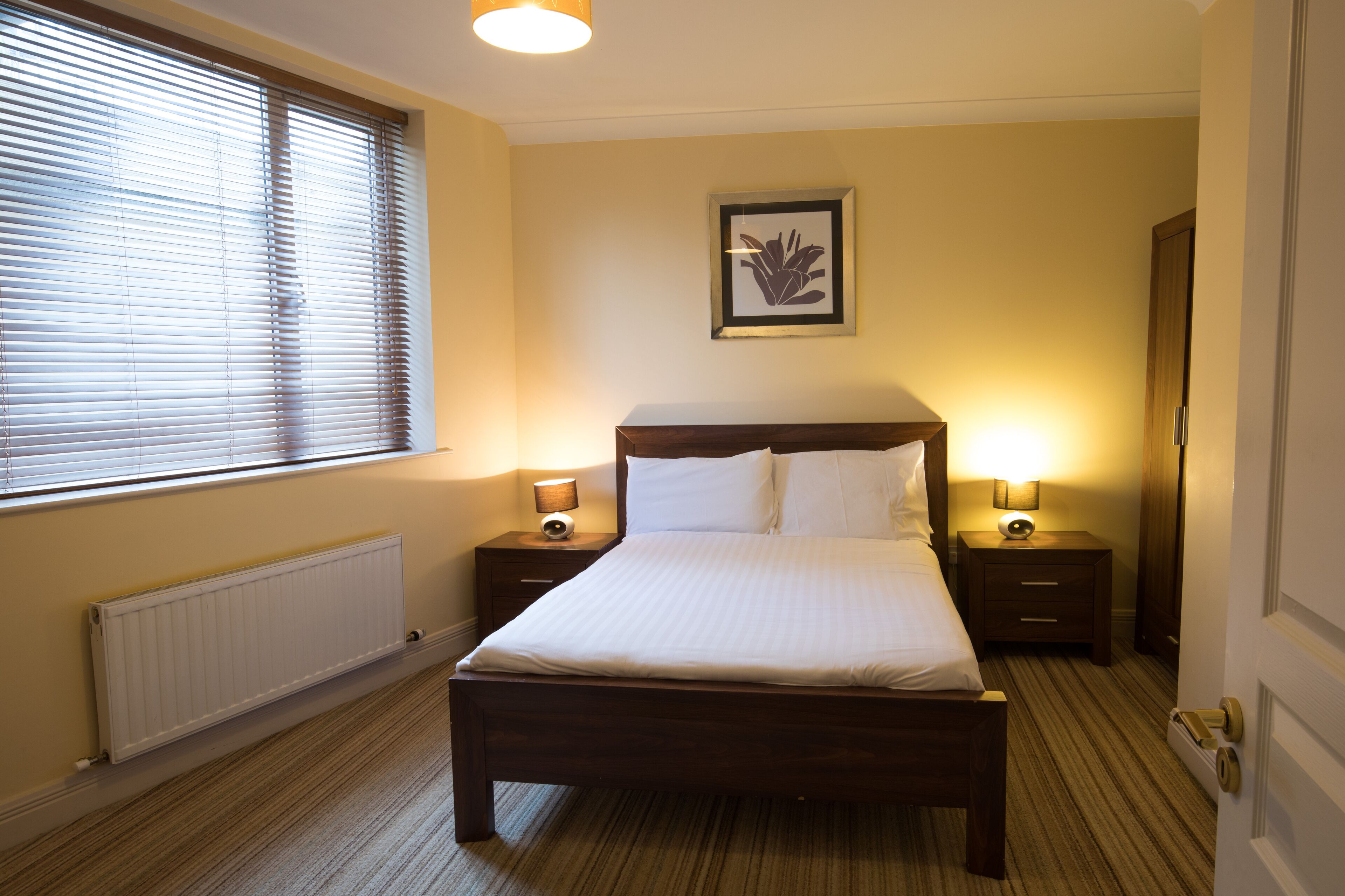 A hotel room with a large bed, two bedside tables with lamps, a framed picture on the wall, and a radiator under the window.