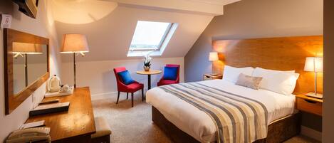 A hotel room with a large bed, a desk with a telephone, two chairs, a small table with a plant, and a skylight.