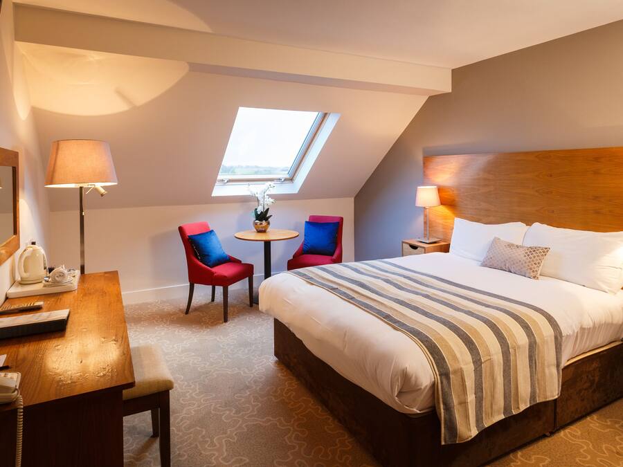 Habitación de hotel con una cama grande, un escritorio con teléfono, dos sillas, una mesita con una planta y un tragaluz.