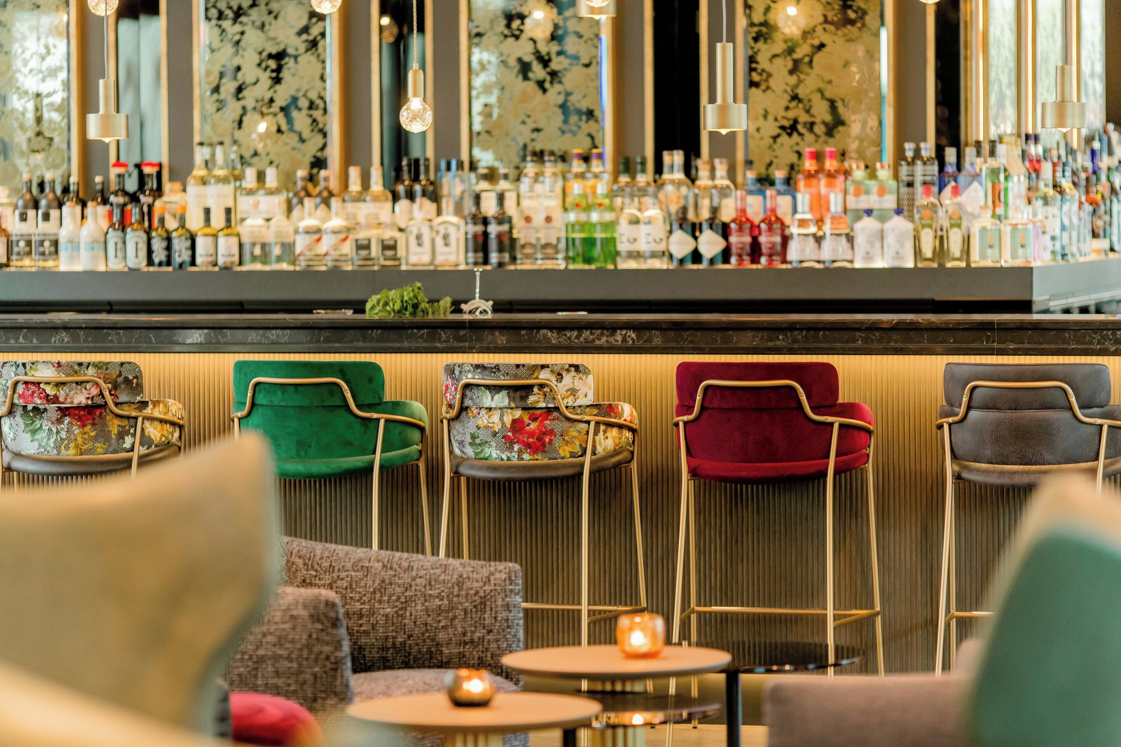 A bar area with a variety of bottles, a green upholstered chair, a red upholstered chair, and a grey upholstered chair.