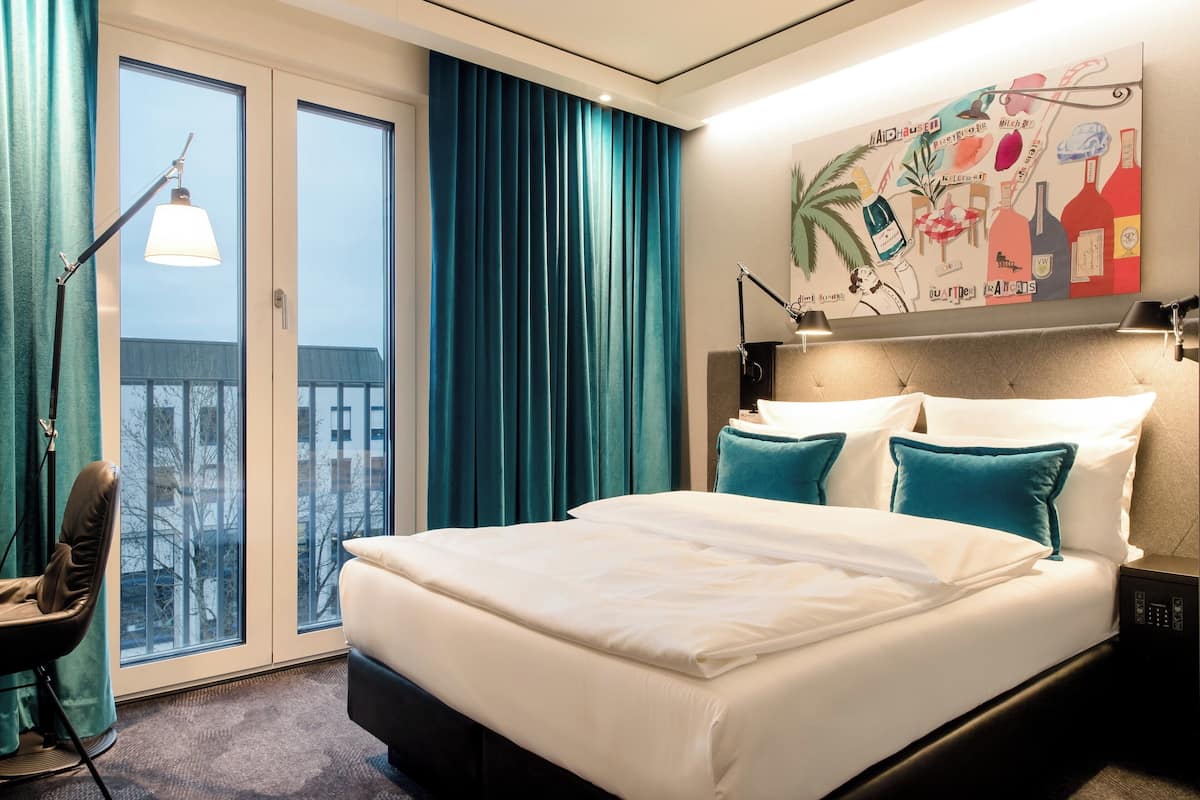 A hotel room with a large bed, a view through the window, a painting on the wall, and a desk lamp.