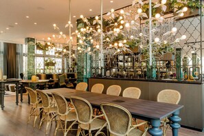 A restaurant with a long wooden table and wicker chairs, a bar area with a green-tiled backsplash, and hanging light fixtures with leaves. - Motel One München-Haidhausen (Munich)