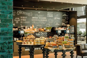 Un café con una pared de pizarra que muestra saludos y artículos del menú. Hay mesas con diferentes platillos y una máquina de café al fondo.
