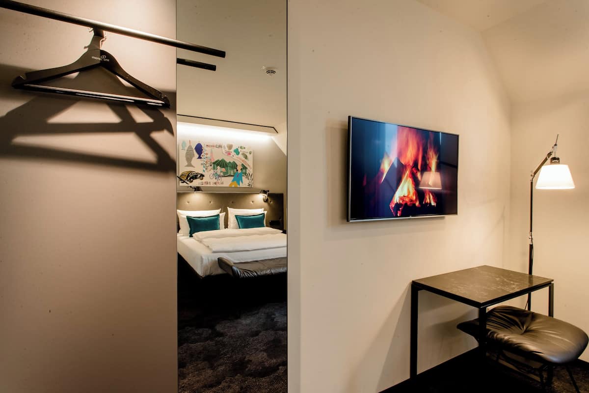 A hotel room with a bed, a desk, a chair, a hanging coat rack, a wall-mounted TV, and a lamp.