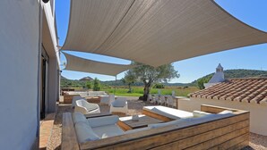 An outdoor seating area with wooden benches and white sofas, shaded by a large canopy, overlooking a scenic landscape with hills and trees. - Tapada do Gramacho (Lagoa)