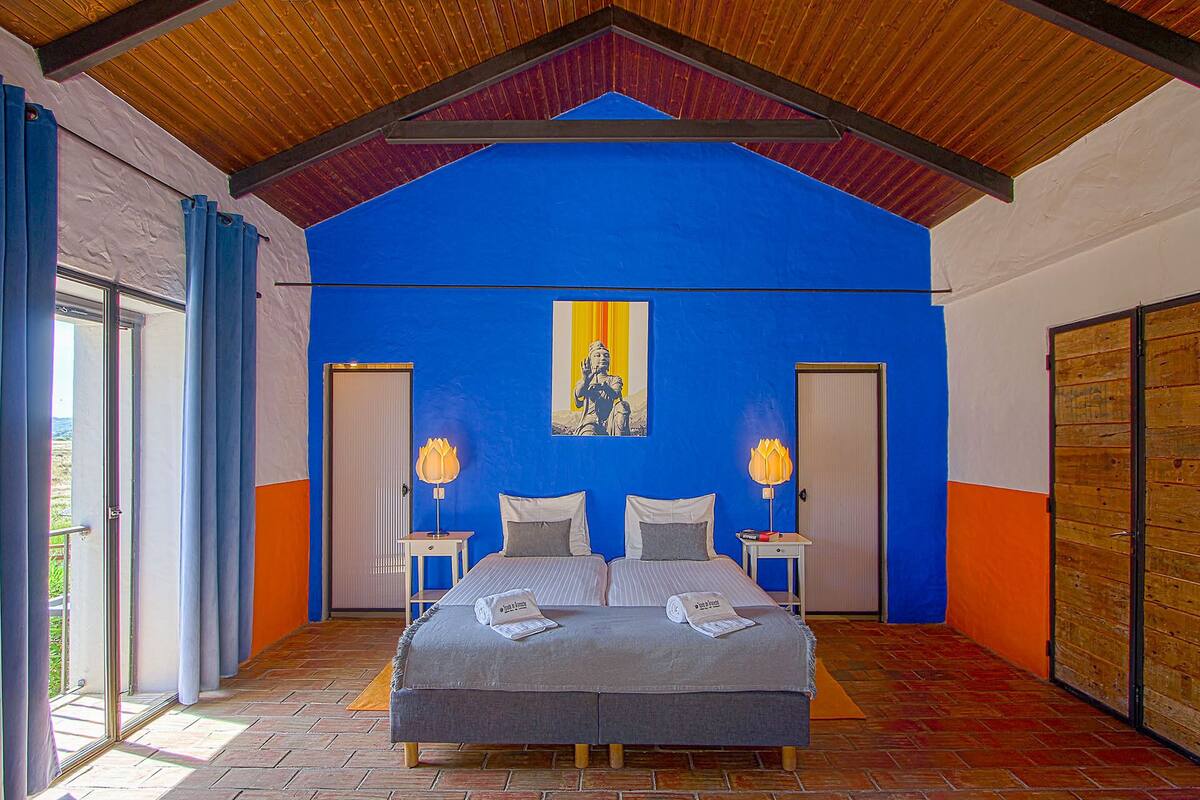 Un dormitorio con una cama grande, dos mesitas de noche, una mesita auxiliar, un ventanal con cortinas y techo de madera.