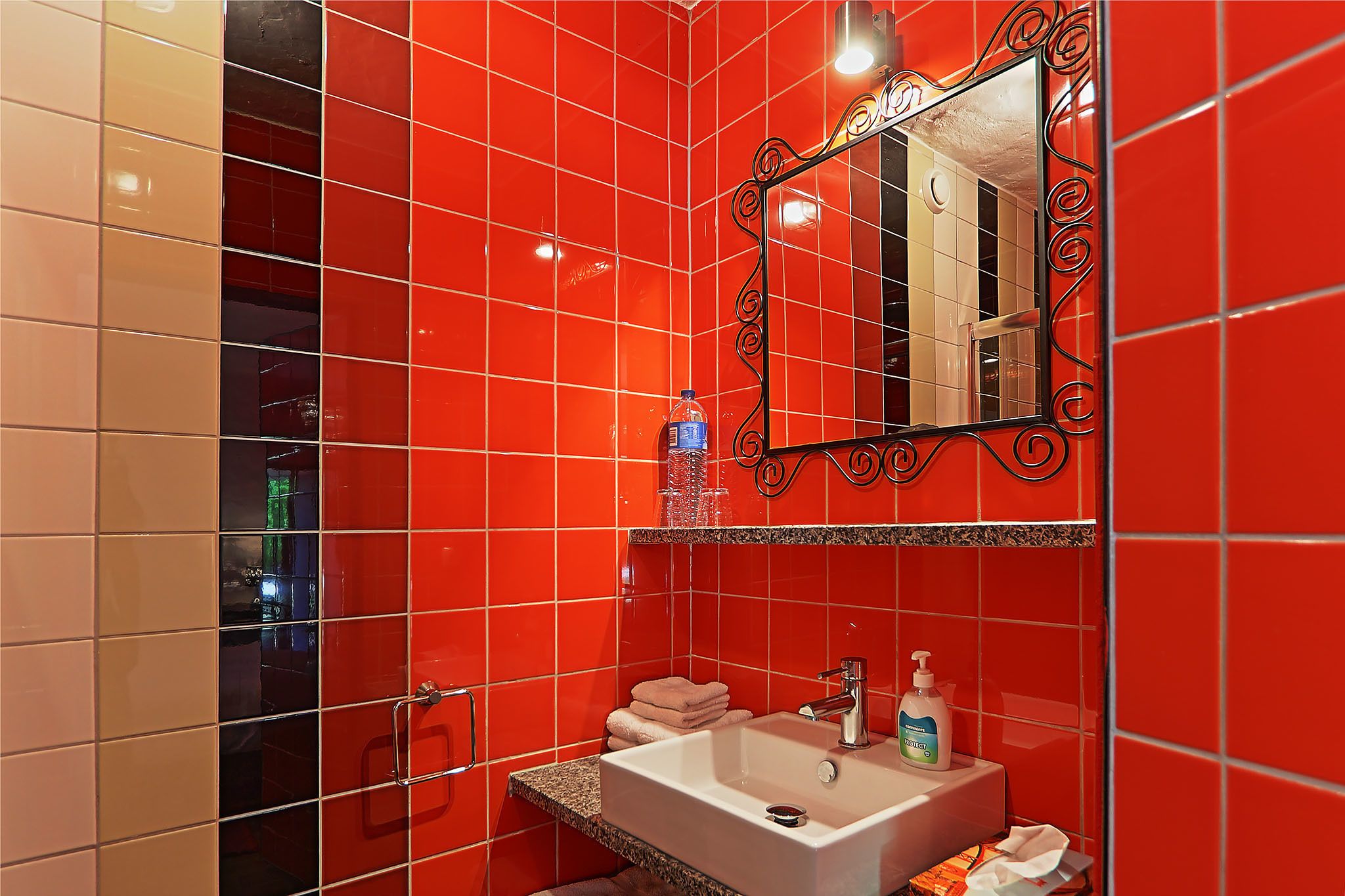 A bathroom with red tiles, a sink with a mirror, and a bottle of hand soap.