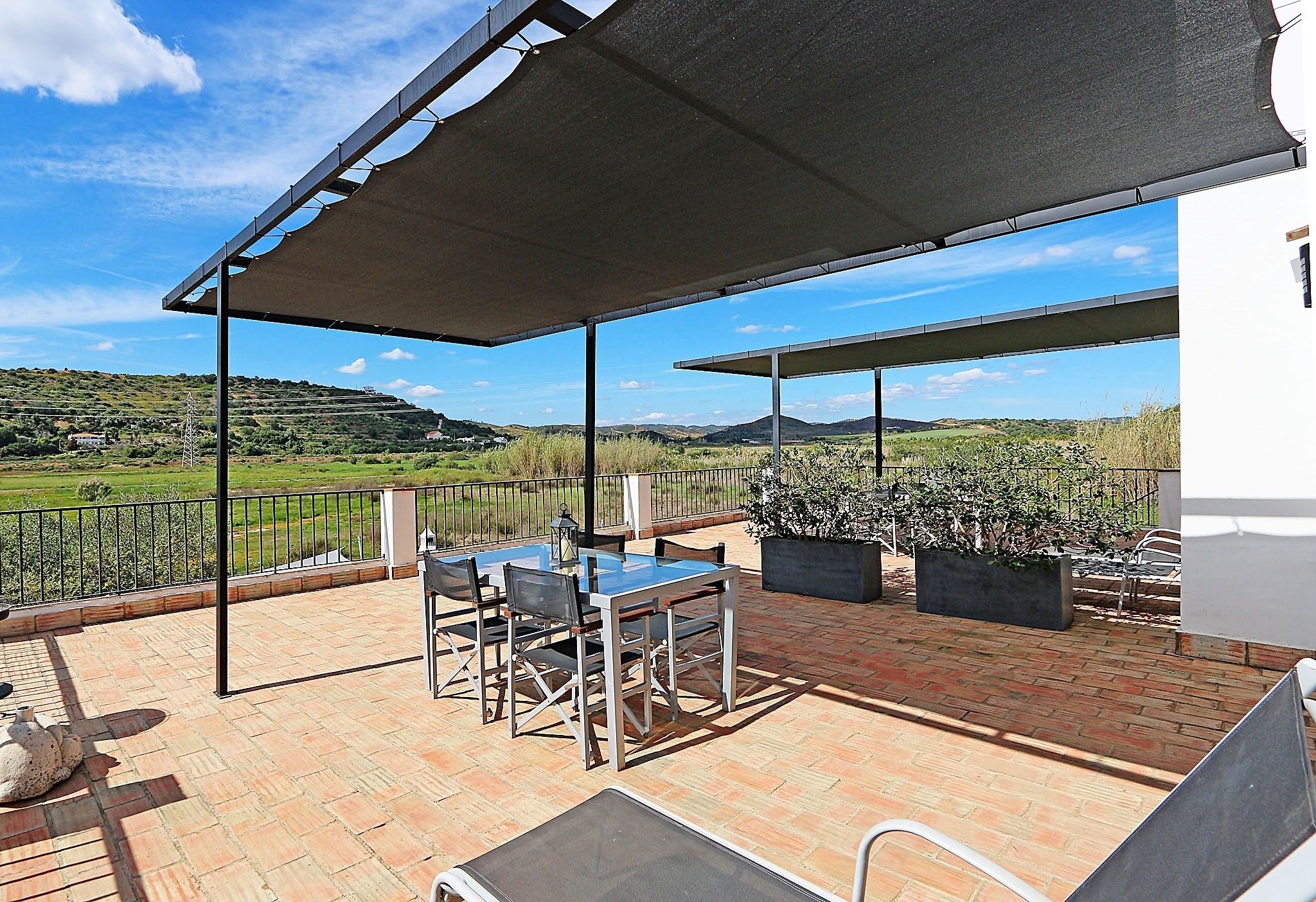 A covered outdoor patio with a table and chairs, a view of the countryside, and a potted plant.