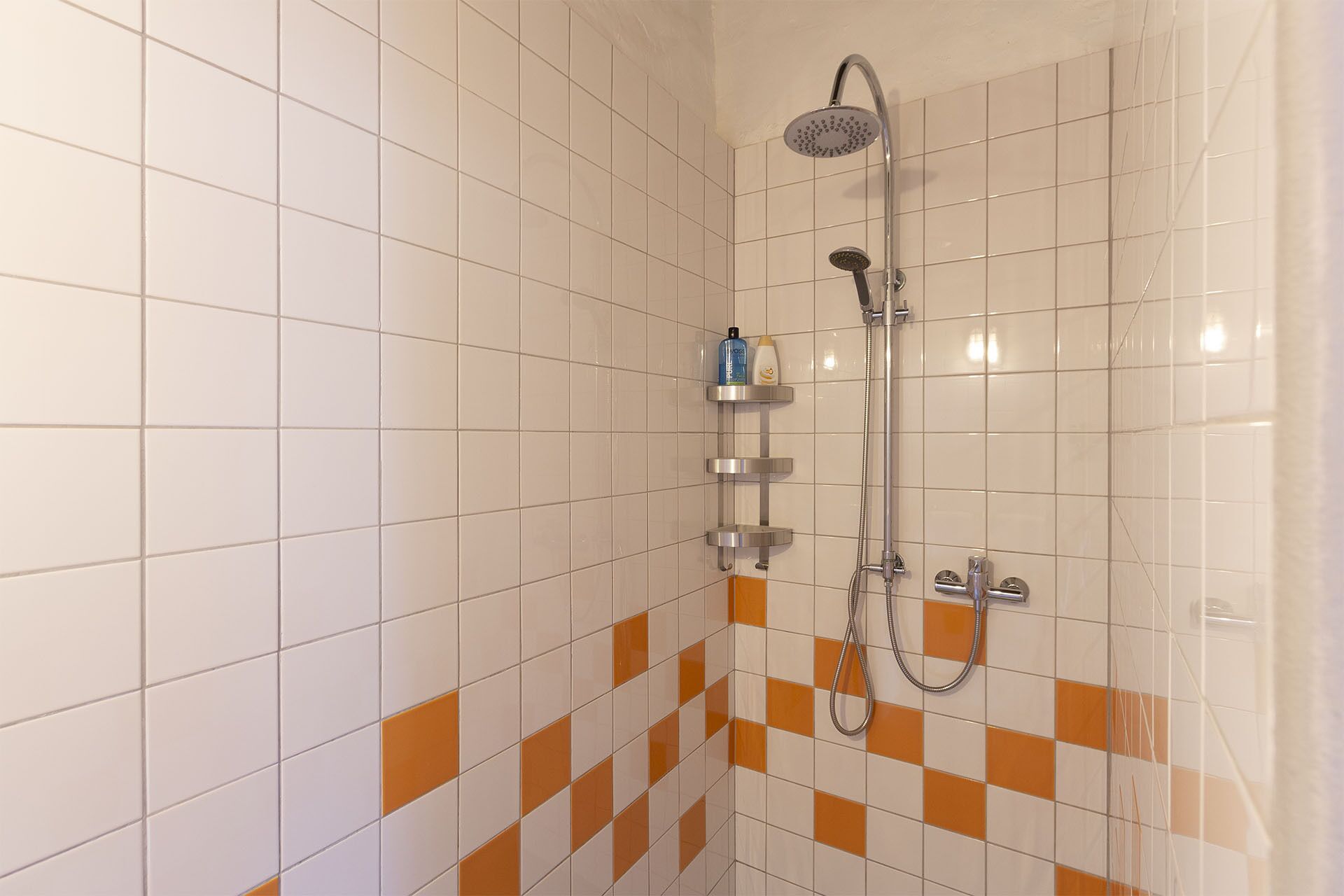 Et brusebad med brusehoved, der kan holdes i hånden, og en vandhane monteret på væggen. To hylder med toiletartikler.