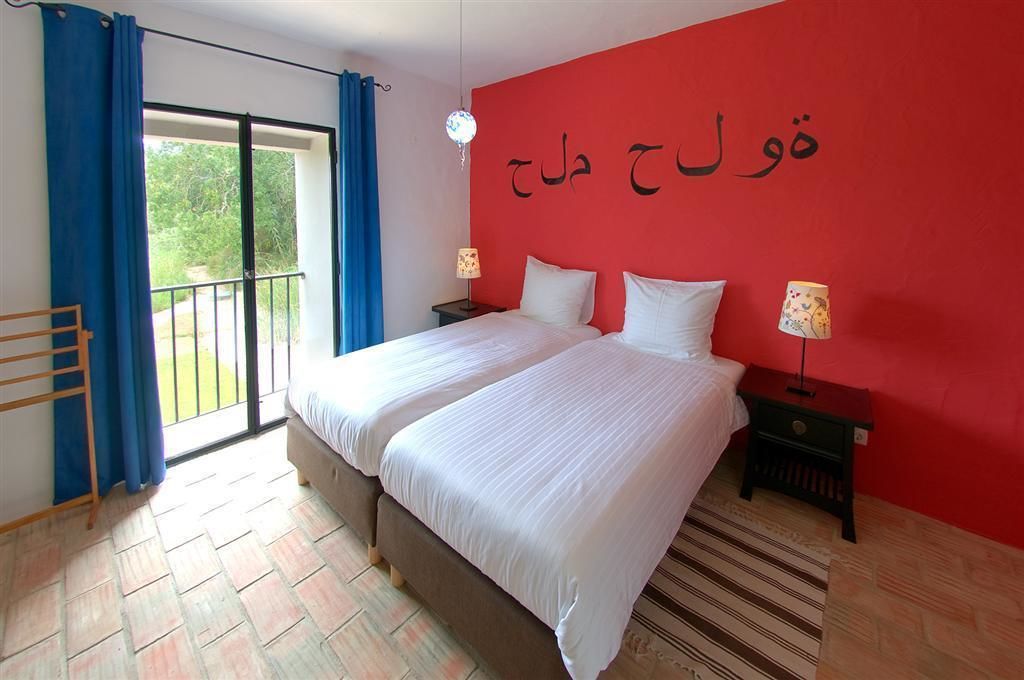 A bedroom with two beds, a red wall, and a balcony with blue curtains.