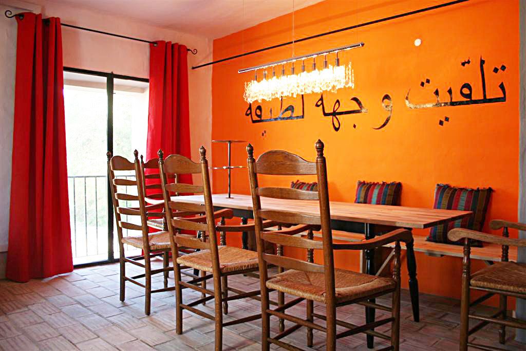 Un coin repas avec une table et des chaises en bois, des rideaux rouges et un mur orange orné d’une écriture arabe.