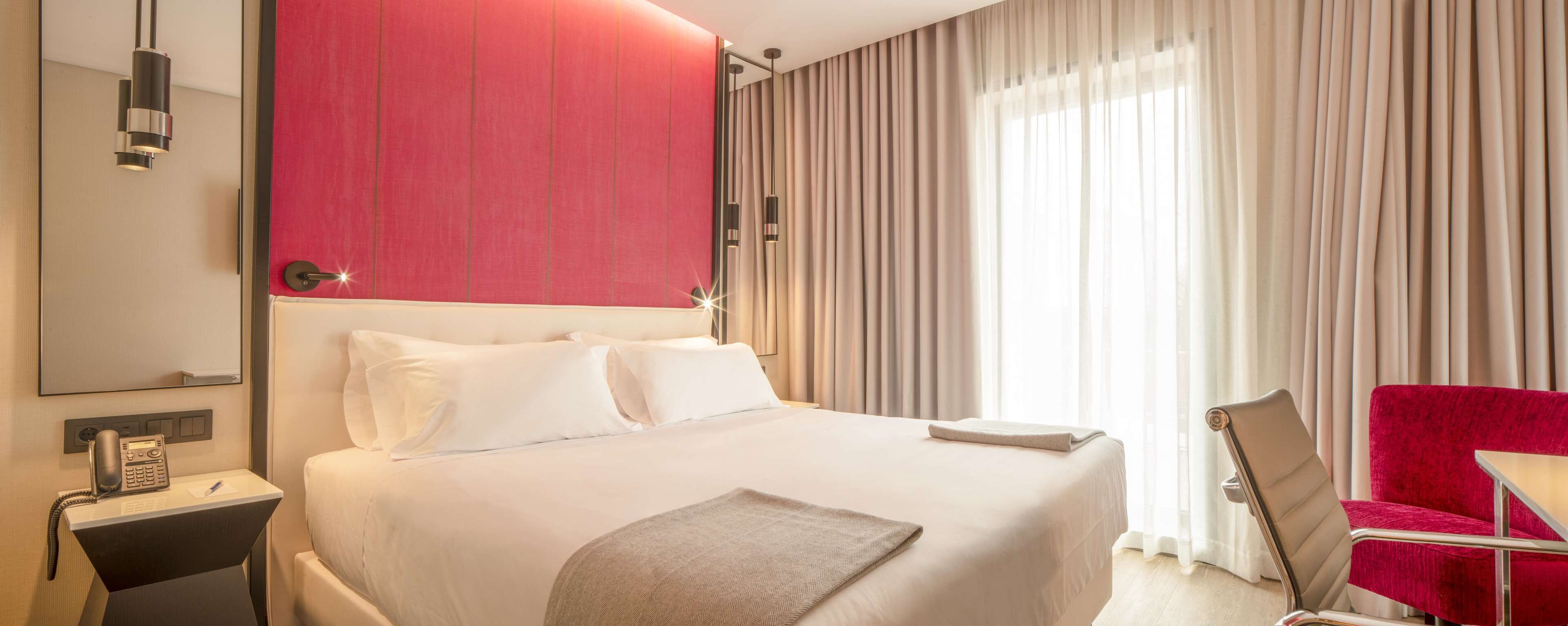 A hotel room with a large bed, a red accent wall, a desk with a phone, a chair, and a window with curtains.
