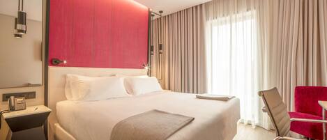 A hotel room with a large bed, a red accent wall, a desk with a phone, a chair, and a window with curtains.