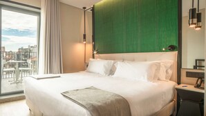 Un cuarto de hotel con una cama grande, un sillón tapizado de color verde, una mesita redonda y vista a la ciudad a través de un ventanal amplio.