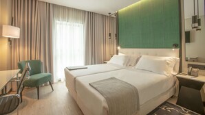 Habitación de hotel con dos camas, un escritorio, una silla y un gran ventanal con cortinas.