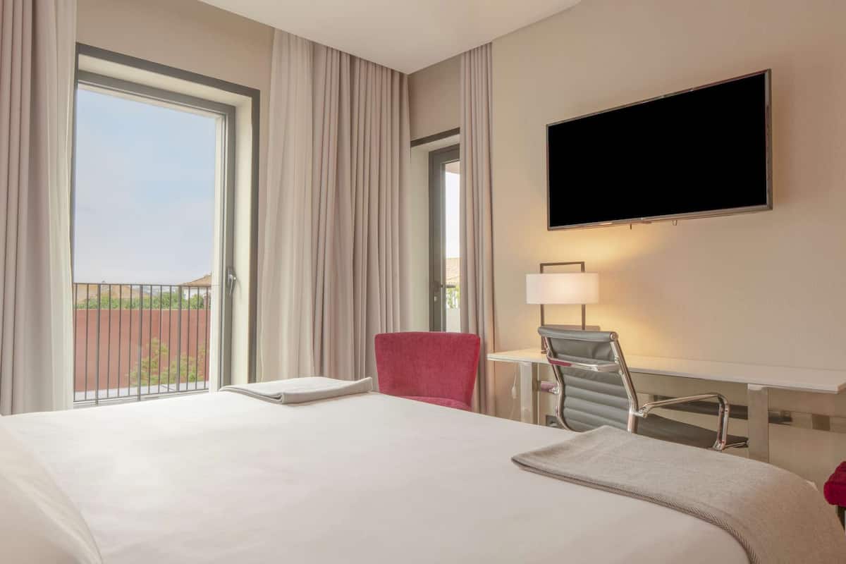 A hotel room with a large bed, a desk with a chair, a flat-screen TV, and a window with curtains.