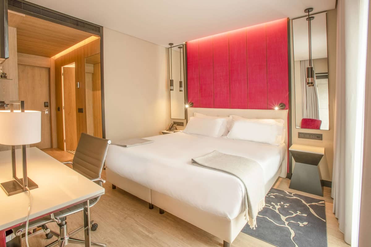 A modern hotel room with a large bed, a desk with a lamp, a chair, and a small table.