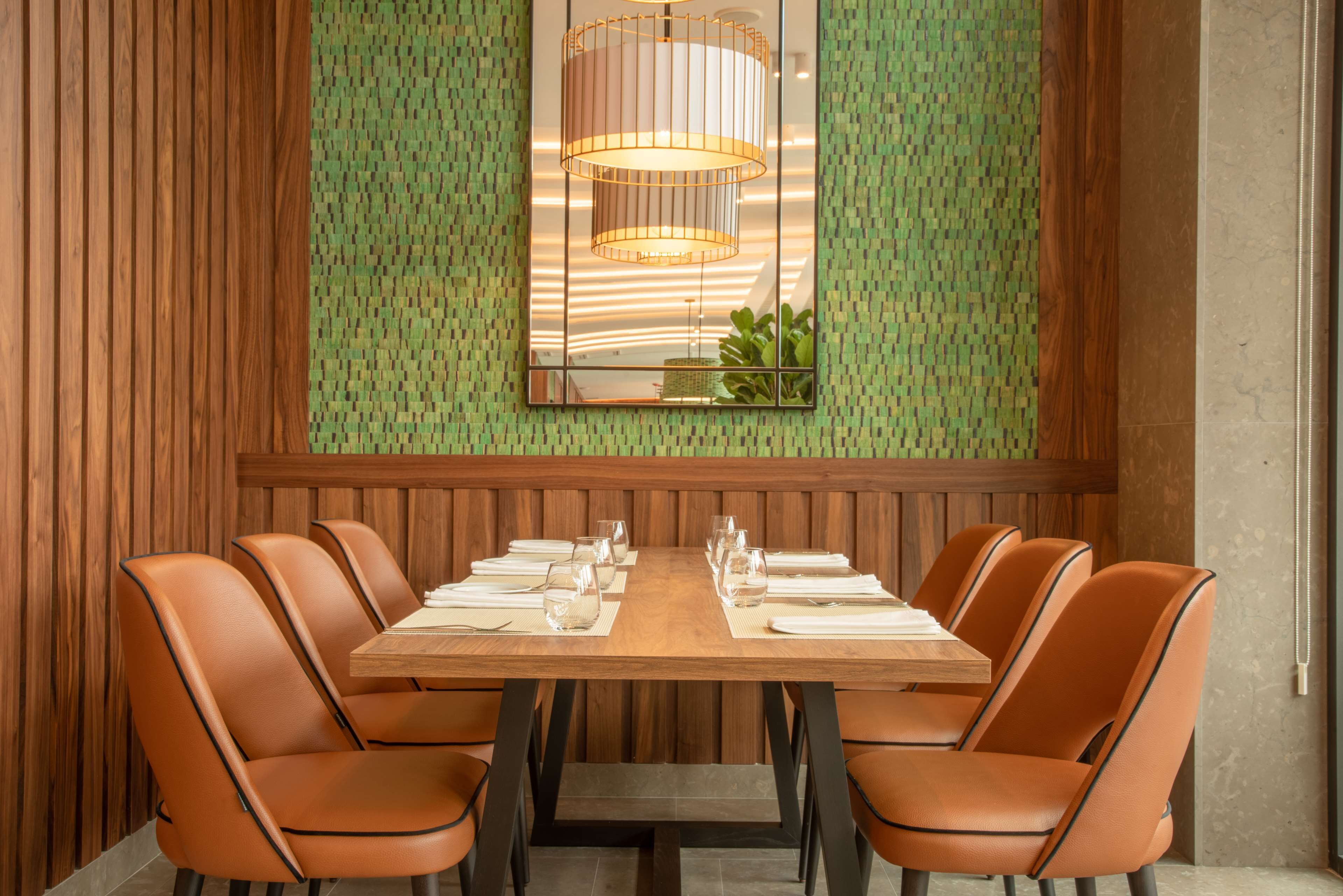 Un comedor con una mesa de madera dispuesta para seis personas, rodeada de sillas de cuero, con una pared verde estampada y una lámpara colgante arriba.