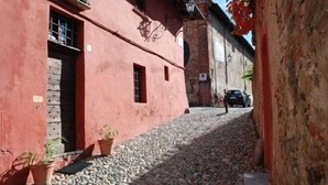 A narrow cobblestone street with buildings painted in a reddish hue. There are potted plants along the sidewalk and a car parked on the street. - B&B San Bernardo (Saluzzo)