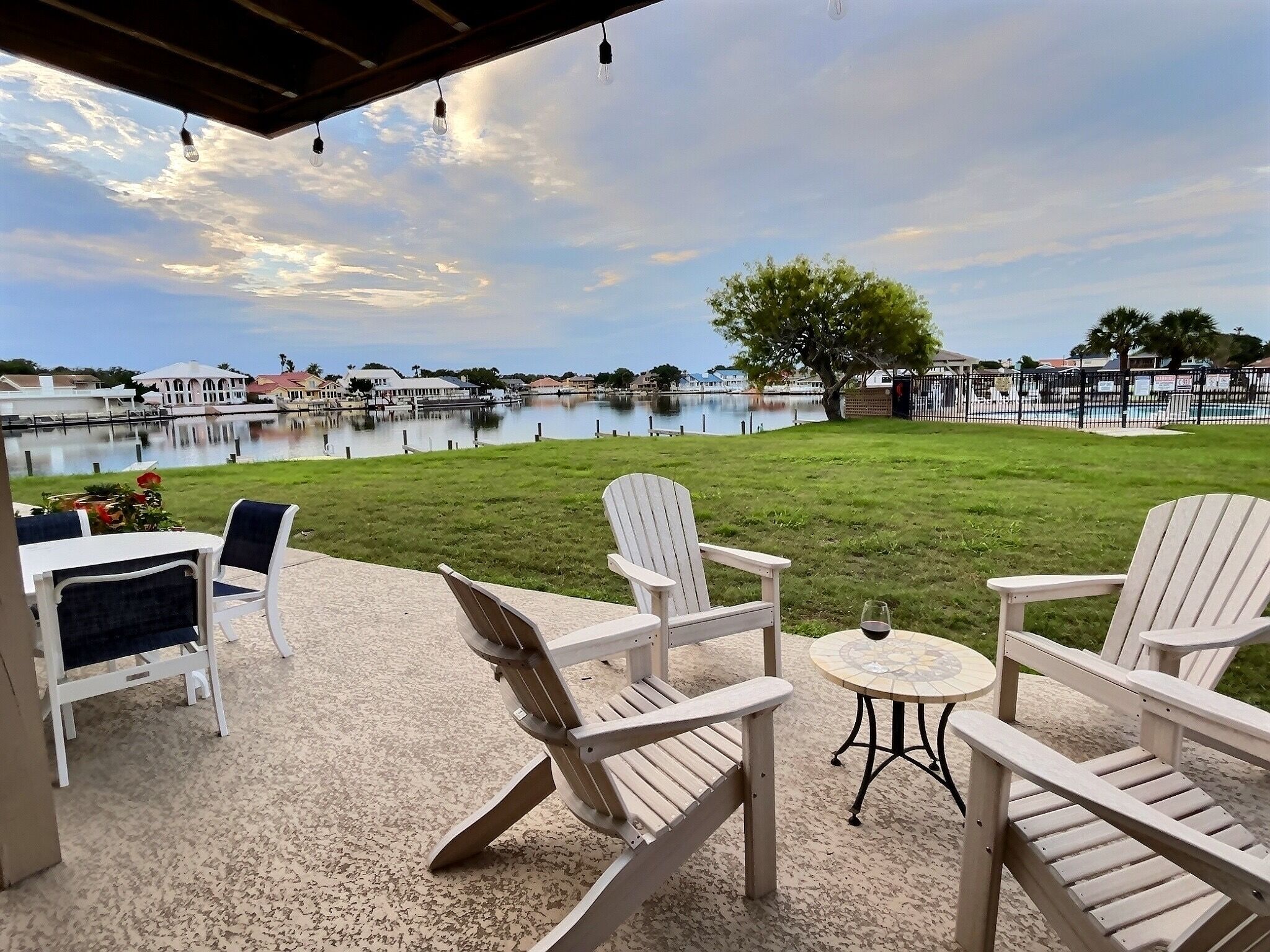 A patio with Adirondack chairs, a small round table, and a view of a lake with a tree and buildings in the background.