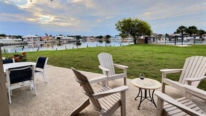 Un patio con poltrone Adirondack, un tavolino rotondo e vista su un lago con un albero e degli edifici sullo sfondo.