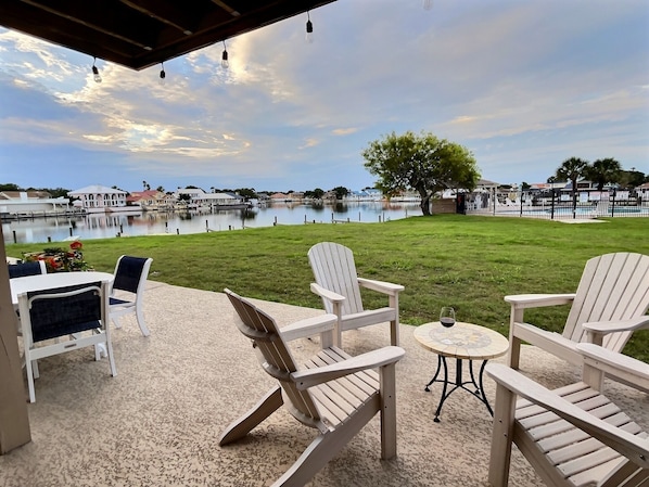 A patio with Adirondack chairs, a small round table, and a view of a lake with a tree and buildings in the background.