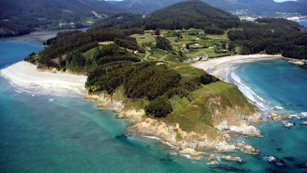 An aerial view of a coastal area with a beach, forested hills, and a small settlement. - Hostal Restaurante La Cepa (Carino)