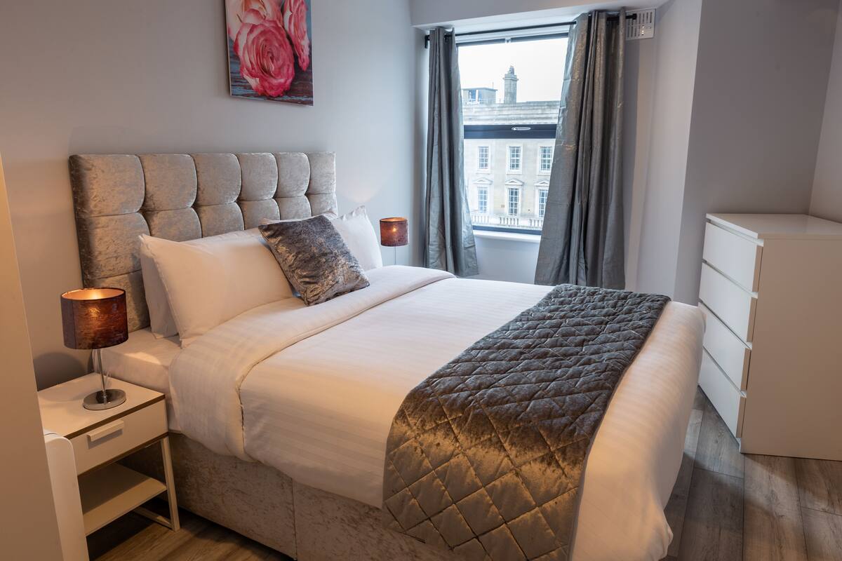 A modern hotel room with a large bed, a nightstand with a lamp, a dresser, and a view of a building through the window.