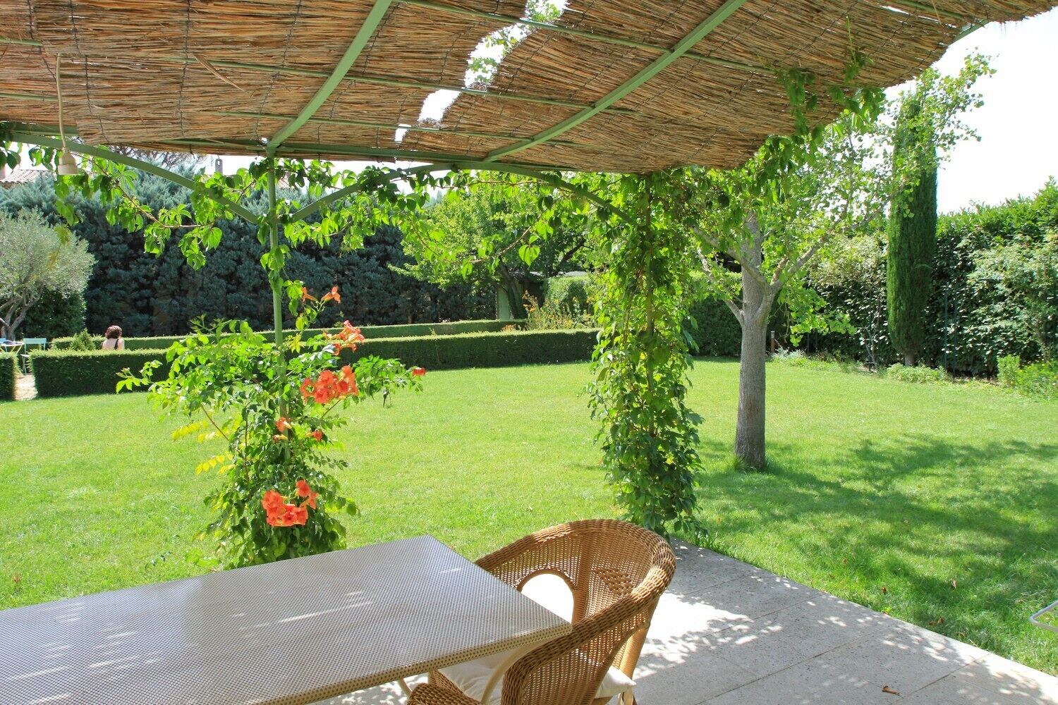 An outdoor patio with a table and wicker chairs under a thatched roof, surrounded by a well-maintained garden and hedge.