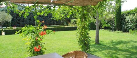 An outdoor patio with a table and wicker chairs under a thatched roof, surrounded by a well-maintained garden and hedge.
