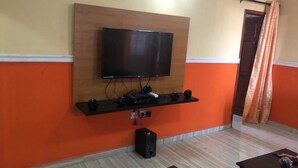 A room with a mounted TV, a wooden shelf with electronic devices, a speaker, and a door with curtains. - Pent House Appartment (Lagos)